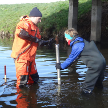 Image captured of technician Uwe Kiwel and researcher Julia Kunz use a metal pipe and hammer to insert a miniDOT logger beneath the streambed.